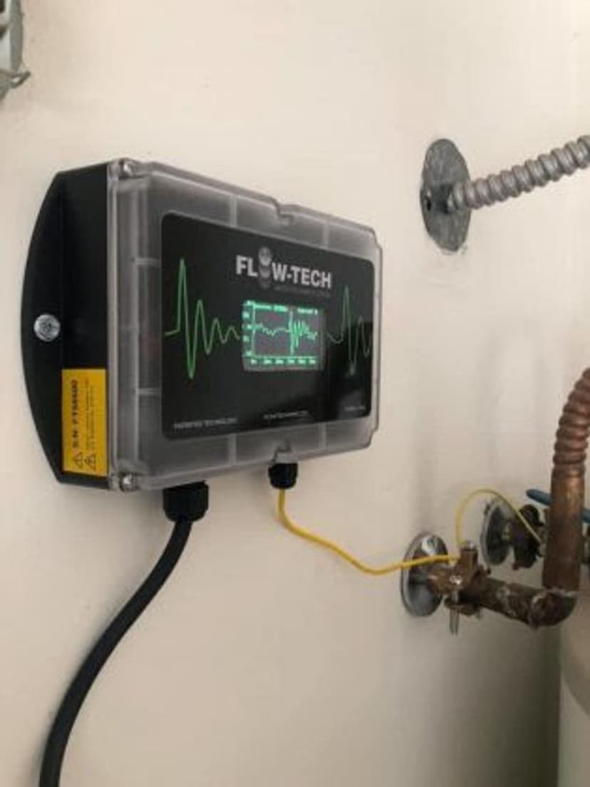 Flow-Tech device mounted on a wall for monitoring water flow, featuring a display screen and connections.
